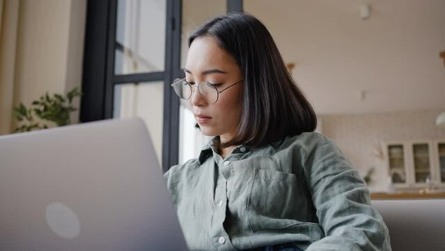 Young Asian woman works on laptop sitting in light apartment