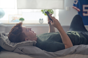 Caucasian teenage boy browsing phone while lying on bed on his back
