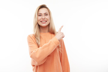 Joyful blonde girl in orange casual smiling points finger at empty copy space for text or product isolated on white background, advertising banner