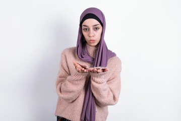 muslim woman wearing hijab and knitted sweater over white background sending blow kiss with pout lips and holding palms to send air kiss.