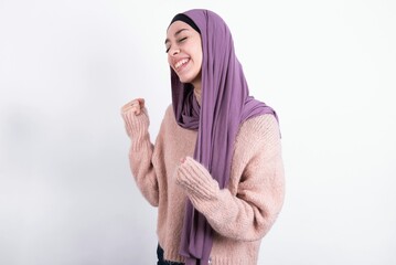 beautiful muslim woman wearing hijab and warm jumper over white background very happy and excited doing winner gesture with arms raised, smiling and screaming for success. Celebration concept.