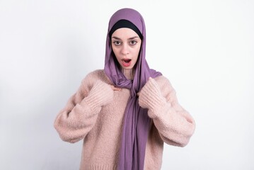 Embarrassed beautiful muslim woman wearing hijab and warm jumper indicates at herself with puzzled expression, being shocked to be chosen to participate in competition, hesitates about something