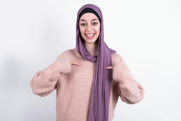 beautiful muslim woman wearing hijab and warm jumper over white background points at his body, being in good mood after going shopping and making successful purchases