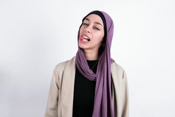 Funny young beautiful muslim woman wearing hijab and jacket over white background makes grimace and crosses eyes plays fool has fun alone sticks out tongue.