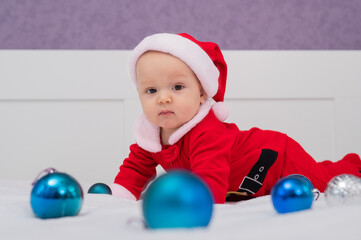 Fototapeta na wymiar Baby boy dressed as Santa is playing with Christmas decorations on the bed.