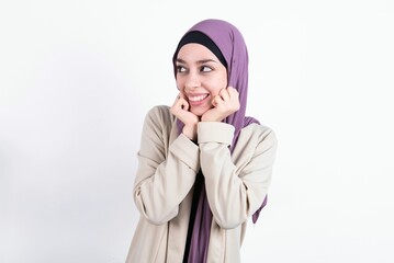 Happy young beautiful muslim woman wearing hijab and jacket over white background anticipates something awesome happen, looks happily aside, keeps hands together near face, has glad expression.