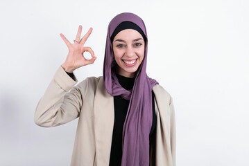 young beautiful muslim woman wearing hijab and jacket over white background hold hand arm okey symbol toothy approve advising novelty news