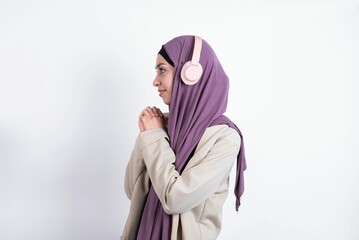 young beautiful muslim woman wearing hijab and jacket over white background wears stereo headphones listening to music concentrated and looking aside with interest.