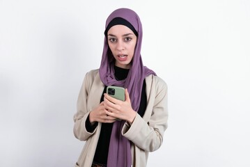Photo of interested young beautiful muslim woman wearing hijab and jacket over white background  hold telephone look side empty space screen