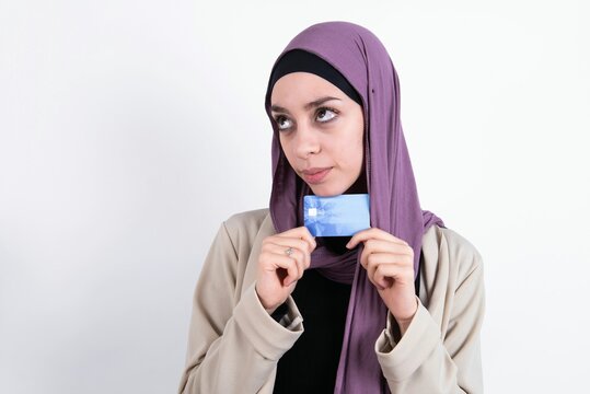 Photo of cheerful young beautiful muslim woman wearing hijab and jacket over white background hold debit card look empty space