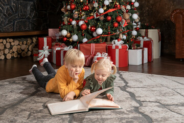 Siblings children on Christmas Eve. Little fair-haired brother and sister read book near the...