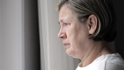 Depressed mature woman cries standing near window in bedroom. Female person wipes tears feeling heartache after loosing loved relative closeup