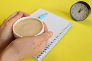 white coffee cup in female hands, yellow background, black alarm clock, morning coffee