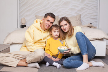 Friendly cute family, parents in yellow clothes with child son sitting by the bed and eating popcorn, looking to the camera