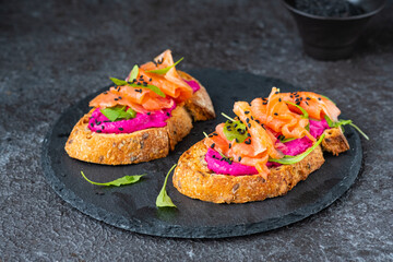 Two open sandwiches with beetroot puree, arugula and salted salmon on a black ceramic board against a dark beon background. Recipes for sandwiches