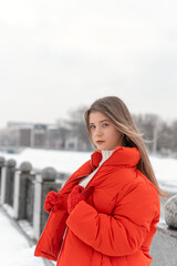 Obraz premium Portrait of beautiful girl in unbuttoned red down jacket and knitted mittens on winter bridge. Warm outerwear for women