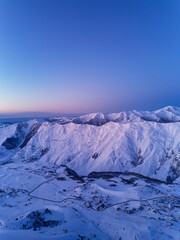 Aerial of snowy mountain range on winter sunrise at ski resort. Mountains valley and village with switchbacks road at sunset. Caucasus peaks skyline in the dusk. City lights at night.