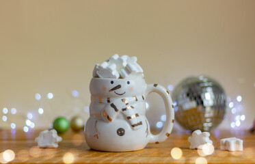 a cup in the shape of a snowman with a white marshmallow on the table on the background of festive lights