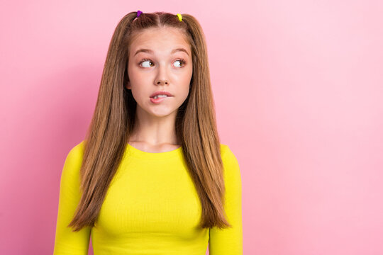 Photo of positive schoolgirl with ponytails dressed yellow long sleeve look empty space biting lip isolated on yellow color background