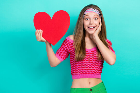 Photo of sweet impressed schoolgirl wear pink crop top rising red paper heart arm hand cheek cheekbone isolated turquoise color background
