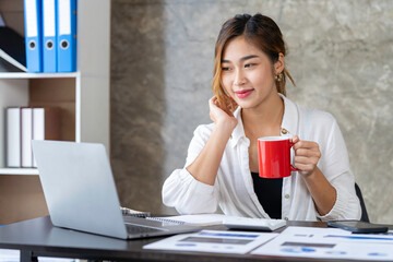 Attractive businesswoman holding cup of hot drink and looking away with smile while sitting in office.