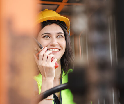 Female foreman using radio while driving forklift vehicle at shipping container yard. Industrial engineer woman drives reach stacker truck at freight cargo warehouse port and use radio communication.