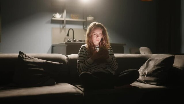 Woman scrolling smartphone, web surfing, sitting on sofa at stylish living room at night. Female playing mobile games or shopping in store. Girl with phone enjoys chatting in social networks relaxing.