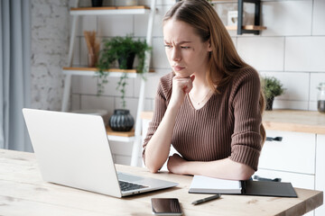 Serious caucasian young female remote worker in casual outfit sitting at table with smartphone and...
