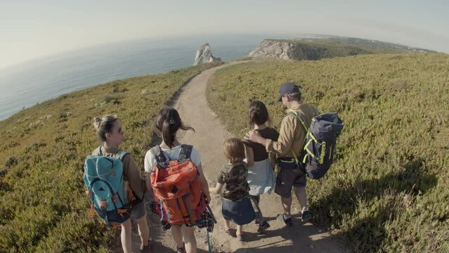 Caucasian family backpacking together, hiking in mountains. Back view of parents and children walking along path towards the sea. Fish eye, crane shot. Slow motion. Tourism, family concept.