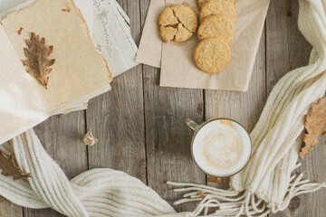 On the wooden table there is a mug of hot coffee with foam, cookies, a book, a branch of mountain ash and a warm scarf. The concept of warmth, comfort