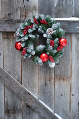 Red balls and snow-covered cones on artificial fir green wreath on Christmas eve. Snowy cozy winter in countryside. Vintage holiday decoration with red holly berries on old door made wooden planks.