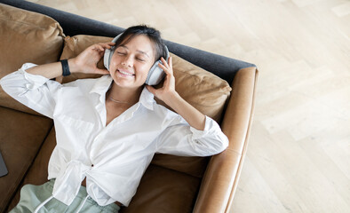 Smiling, listening to an audiobook with headphones, using an application with books. A woman is sitting on a leather sofa.