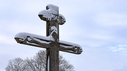 Orthodox wooden cross with a crucifix, sprinkled with snow, against the background of a gloomy winter sky on the street near a road or village, part of the Christian cross. The concept of Orthodoxy.