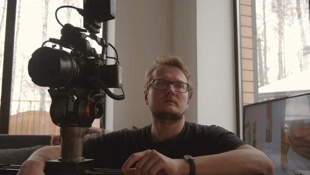 Shoulder-up of young man in eyeglasses filming something using professional video camera indoors at daytime