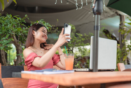 A pretty Asian lady takes a selfie using the front camera of her cellphone as she unwinds in an al fresco cafe with a cold drink.