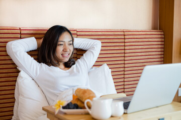 Young Asian women smiling happily at freelance work, working on a notebook while relaxing in bed with snacks and fruit in a hotel room. Vacation and relaxation