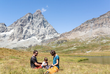 Fototapeta premium Toddler drinking water from a hydration bag while is resting with her parents on a green meadow under the Matterhorn at italian alps.