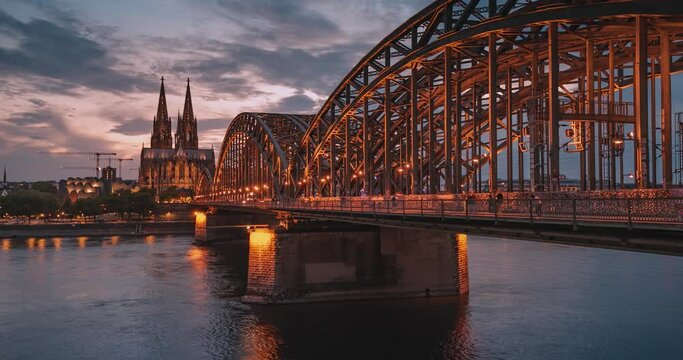 Timelapse video of the illuminated bridge over which trains pass and tourists pass and the Cologne Cathedral in the evening at the blue hour