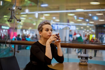A young business woman in a black dress drinks coffee in a cafe, enjoying solitude and thinking about the future. Rest from work