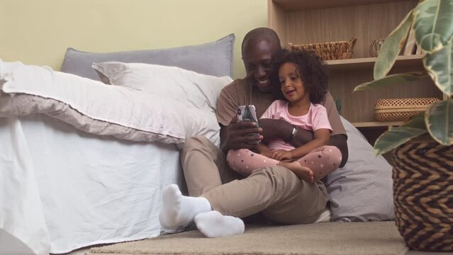 Cheerful Black man with 3 year old daughter video calling mom on smartphone from home