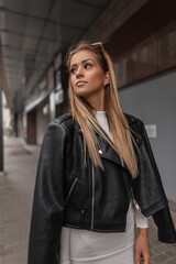 Trendy beautiful young fashion woman model in casual rock black leather jacket and white dress walks on the street on urban background