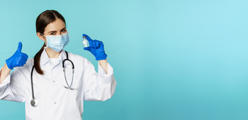 Portrait of smiling doctor, medical personel in face medical mask and rubber gloves, showing thumbs up and coronavirus, omicron vaccine, standing over blue background - obrazy, fototapety, plakaty