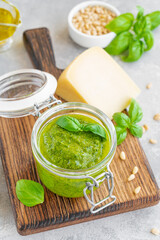 Traditional Italian basil pesto sauce in a glass jar on a board with ingredients for cooking on a gray concrete background. Copy space.