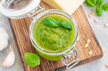 Traditional Italian basil pesto sauce in a glass jar on a board with ingredients for cooking on a gray concrete background. Copy space.