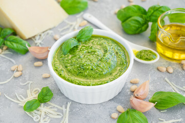 Traditional Italian basil pesto sauce in a white bowl with ingredients for cooking on a gray concrete background. Copy space.