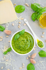 Traditional Italian basil pesto sauce in a white bowl with ingredients for cooking on a gray concrete background. Copy space.