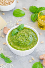 Traditional Italian basil pesto sauce in a white bowl with ingredients for cooking on a gray concrete background. Copy space.