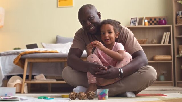Portrait of cheerful African American man and his 3 year old daughter sitting on rug in cozy apartment smiling at camera, drawing and playing together
