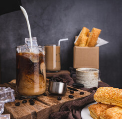 Pouring milk to make latte coffee into a transparent glass with ice. and roasted coffee beans and Crispy Pie dessert on wood and black background studio photo