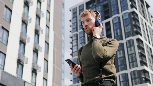 Cheerful fashion guy wearing headphones standing outdoors dancing using smartphone music new app in modern city on summer day. Handsome man having fun, rest on urban street. People and music concept.